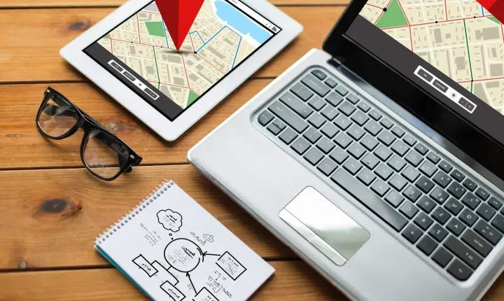 Laptop and tablet screens showing map routes with red location markers on a wooden desk.