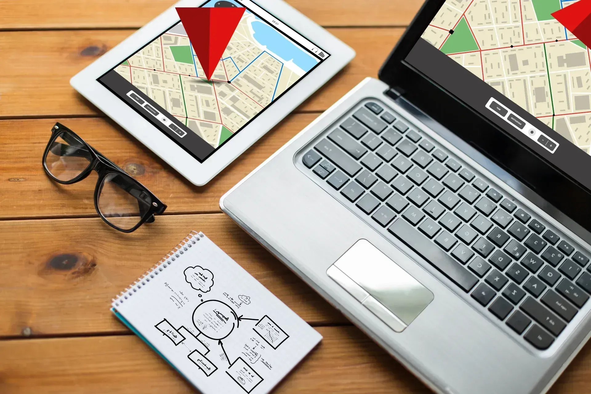 Laptop and tablet screens showing map routes with red location markers on a wooden desk.